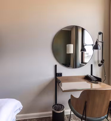 Moderne hotelværelseshjørne med et minimalistisk skrivebord og et rundt spejl over det. En væghængt læselampe og en telefon sidder på skrivebordet, som er parret med en træstol. Et fladskærms-tv er til venstre, og et vindue med gardiner er til højre.