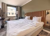 Et moderne hotelværelse har en stor, pænt redt seng med hvidt sengetøj og en polstret sengegavl. Et skrivebord med en stol er nær vinduet, og et lille bord med en stol er på den modsatte side. Væggene er malet grønne.