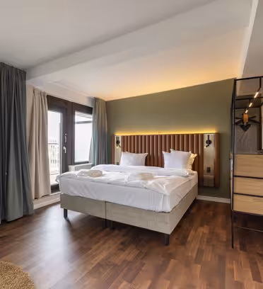 Et moderne hotelværelse med en pænt lavet dobbeltseng med hvidt sengetøj. Værelset har trægulv, tv på væggen, høj lampe og store vinduer med gardiner. Der er et lille rundt bord og en stilfuld reol ved siden af sengen.