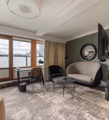 Moderne hotelværelse med store vinduer, beige og grønne vægge og gulvtæppe. Den har en sofa, et rundt spejl, et monteret tv, et lille skrivebord med en stol og et cirkulært sofabord. Et beige gardin dækker delvist vinduet.