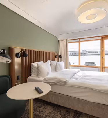 Et hotelværelse med en pænt lavet dobbeltseng. Rummet har en grøn accentvæg og et rundt bord med fjernbetjening til fjernsynet. En plys stol sidder i nærheden. Store vinduespartier giver naturligt lys og udsigt over bybilledet.