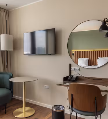 Moderne hotelværelse interiør med et rundt spejl over et skrivebord, en vægmonteret lampe, et tv og en stol. Til venstre et rundt bord med en grøn stol og en gulvlampe ved vinduet, som har beige gardiner. En skraldespand er placeret ved siden af skrivebordet.