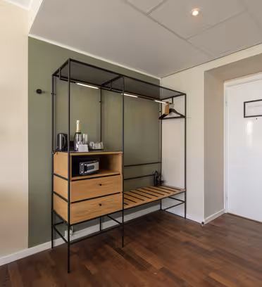 Et moderne hotelværelseshjørne med et elegant åbent klædeskab med hylder og hængende plads. En champagneflaske og glas står på hylden. Et rundt spejl og væglampe er til venstre. Mørkt trægulv og en hvid dør er synlige.