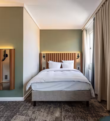 Hotelværelse med to enkeltsenge, hver med en polstret sengegavl og hvidt sengetøj. Der er en lysegrøn accentvæg og beige gardiner ved vinduet. En mørkegrøn stol er delvist synlig i hjørnet. Gulvet har et mønstret tæppe.
