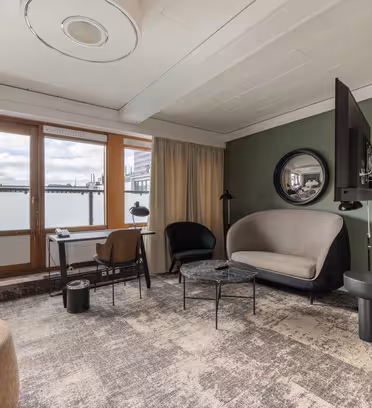 Moderne stue med stort vinduesparti og altanudsigt. Har en beige sofa, grøn lænestol og en sort sidestol omkring et rundt sofabord. Et vægmonteret tv vender ud mod opholdsområdet. Neutrale toner og blød belysning skaber en hyggelig atmosfære.