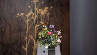 En farverig buket blomster i en vase, placeret foran en væg med et mørkt blomstermønster. To grå stole er delvist synlige bag arrangementet. Rammerne fremstår moderne og elegante.