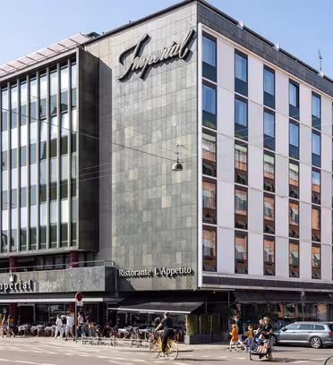En moderne hjørnebygning med store vinduer huser Imperial-hotellet og restauranten LApetito. Folk går og cykler på gaden foran, og biler er parkeret langs vejkanten. Det er en solskinsdag.