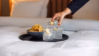 En hånd hælder en klar drink fra en lille flaske op i et glas med is og citron på en bakke, ved siden af et andet glas og en skål chips, alt sammen placeret på en pænt redt seng.