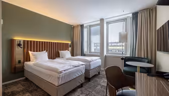 Moderne hotelværelse med to enkeltsenge, grøn accentvæg, store vinduer med udsigt over byen, skrivebord, stole og neutral indretning. Værelset er rent og godt oplyst med naturligt og kunstigt lys.