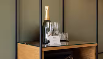 En flaske mousserende vin, to tomme champagneglas og en bakke med indpakkede glas står på en glashylde over et træskab. En velkomsthilsen er placeret foran glassene.
