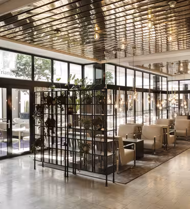 Et moderne lobbyområde med beige lænestole og borde på et klinkegulv. Rummet er opdelt af en metal- og glasskillevæg med planter. Overhead, reflekterende metalpaneler med lys fremhæver den lyse, åbne atmosfære.