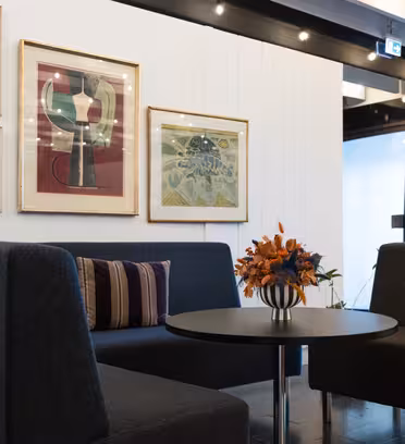 Et moderne opholdsområde med to mørkegrå stole, et rundt sort bord og en vase med orange blomster. Bag dem har en hvid væg flere indrammede abstrakte kunstværker. Rummet har blød belysning og et stilfuldt, moderne præg.