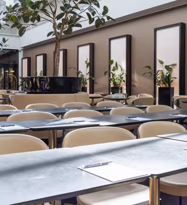 Et moderne mødelokale med rækker af elegante borde og beige stole. Hvert bord har en udklipsholder og pen. Høje planter og store spejle tilføjer indretning. Rummet er godt oplyst med naturligt lys, der kommer gennem vinduerne.