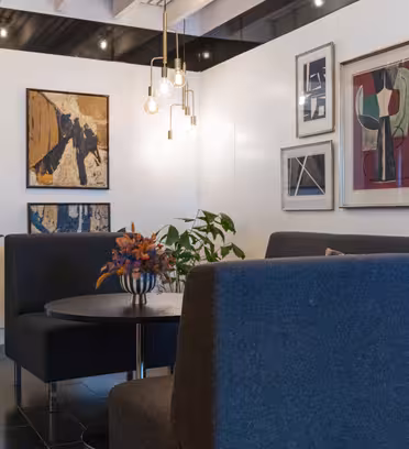Et moderne kunstgalleri med et opholdsområde med blå sofaer omkring et rundt bord prydet med et farverigt blomsterarrangement. Abstrakte malerier dekorerer de hvide vægge, og hængende lysarmaturer giver stemningsbelysning.