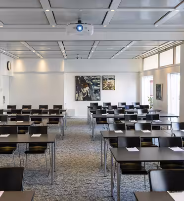 Et mødelokale med rækker af borde og sorte stole indrettet i klasseværelset. Hvert bord har en notesblok og pen. To malerier hænger på væggen, og naturligt lys fylder rummet gennem store vinduespartier. En projektor er monteret på loftet.