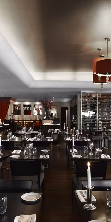 Et moderne restaurantinteriør med mørke træborde, sorte siddepladser og røde accenter. Værelset har en væg af vinflasker, farverige kunstværker, elegante lysekroner og borde dækket med glasvarer og stearinlys.