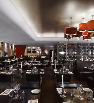 Moderne restaurantinteriør med mørke træborde sat med glasvarer og hvide servietter. Lædersæder, røde lysekroner og farverig vægkunst forbedrer stemningen. En vinreol er synlig i baggrunden.