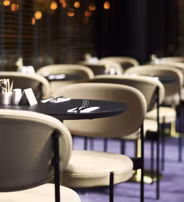 Et moderne restaurantinteriør med rækker af runde borde til spisning. Bordene har sorte overflader og er parret med beige stole. Blød, varm belysning og lodrette persienner skaber en hyggelig atmosfære. Gulvet er belagt med et lilla tæppe.