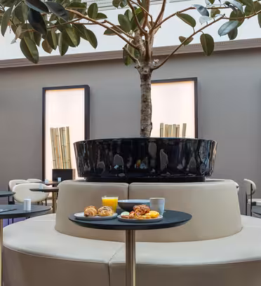 Et moderne caféinteriør med et centralt cirkulært siddeområde med et stort pottetræ. Små borde omgiver siddepladserne, og et bord viser tallerkener med croissanter og frugt sammen med et glas appelsinjuice.