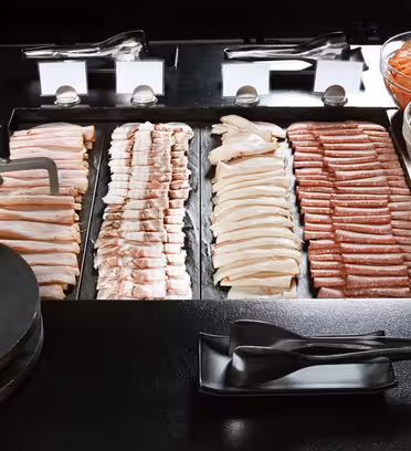 En buffet med et udvalg af råt kød arrangeret i pæne rækker, inklusive baconstrimler og pølselinks. Til højre er skåle med skåret agurker, tomater og peberfrugter. Serveringsredskaber stilles foran.