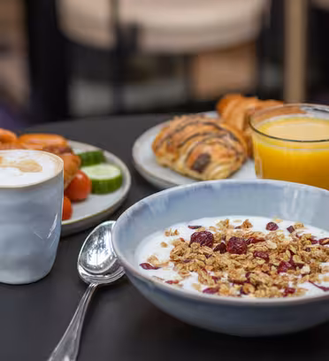 En morgenmadsindstilling med en skål yoghurt toppet med granola og tørrede bær, en kop skummende kaffe, et glas appelsinjuice og en tallerken med croissanter, appelsinskiver og grøntsager på et mørkt bord.