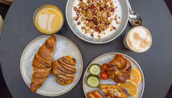 Et bord dækket til morgenmad med en skål yoghurt toppet med granola og tranebær, et glas appelsinjuice, en kop cappuccino, croissanter, en kanel wienerbrød, skiver af appelsin, agurk, tomater, pølser og bacon.