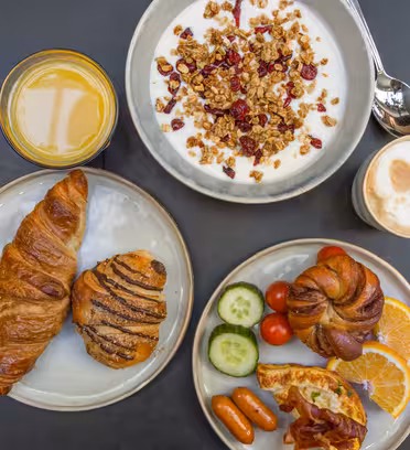 Et bord dækket til morgenmad med en skål yoghurt toppet med granola og tranebær, et glas appelsinjuice, en kop cappuccino, croissanter, en kanel wienerbrød, skiver af appelsin, agurk, tomater, pølser og bacon.