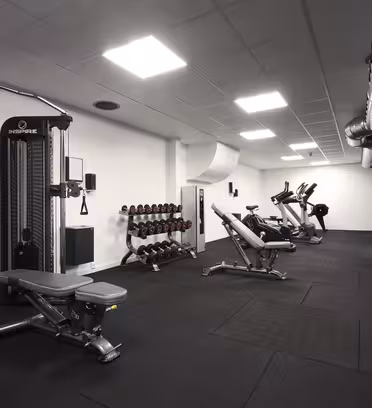 Et moderne fitnesscenter interiør med sorte gulve og hvide vægge. Den har træningsmaskiner, herunder elliptiske, stationære cykler og en multi-gym-station. Håndvægte er pænt arrangeret på stativer. Loftskanaler og belysning er synlige.