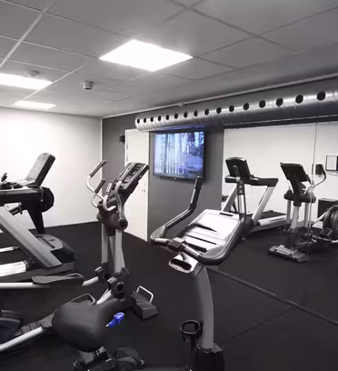 Et moderne fitnessrum med løbebånd og motionscykler. Rummet er rent og slankt med et stort spejl på den ene væg og en metalluftkanal på loftet, hvilket skaber et industrielt look. Rummet er godt oplyst af loftslamper.