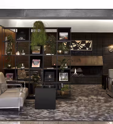 En moderne lounge med grå lænestole, et plys tæppe og en sort reol prydet med planter, bøger og kunst. En pejs er i baggrunden, og træpaneler tilføjer varme. Atmosfæren er hyggelig og stilfuld.